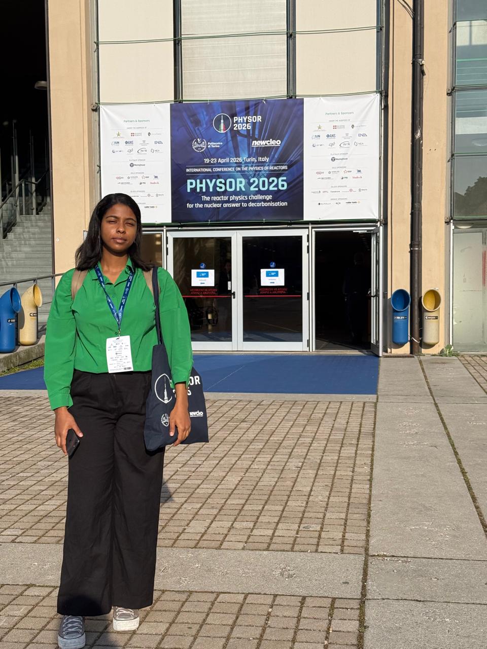 A picture of Dina standing in front of the main enterance to the Centro Congressi Lingotto with a poster for PHYSOR 2026