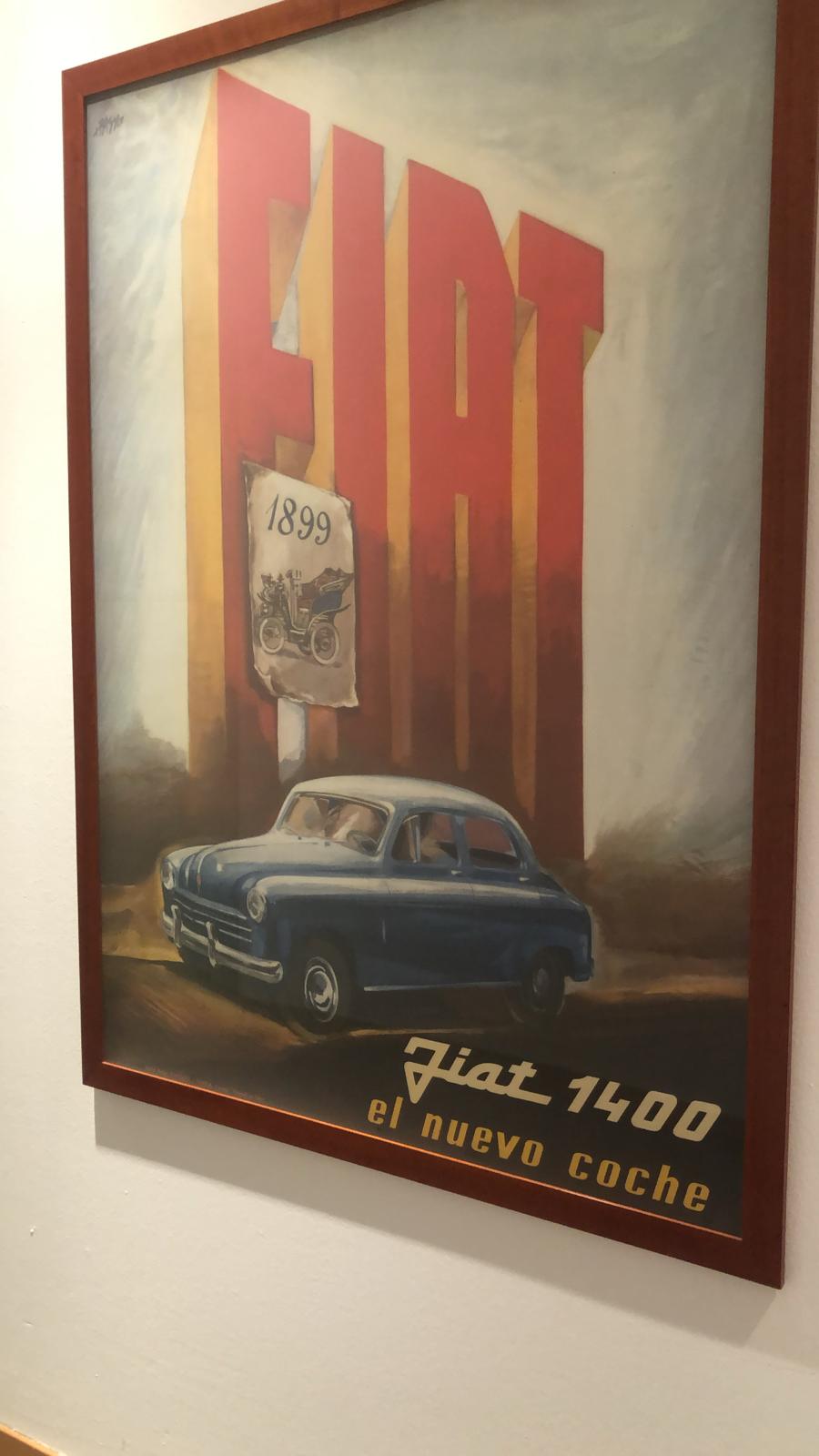 A picture of a FIAT 1400 advertising poster hung at the Centro Congressi Lingotto in Turin, Italy. FIAT In big letters with a smaller poster of their automobiles from 1899 is part of the poster, and the FIAT 1400 driving by. el neuvo coche is written at the bottom right.